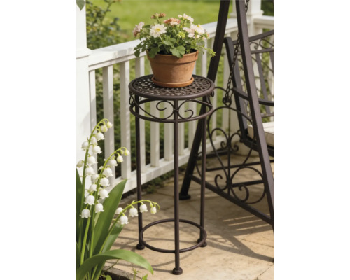 Hoher Blumenständer aus Metall mit Ornamenten auf einer Terrasse neben einer Gartenbank und einem Geländer.