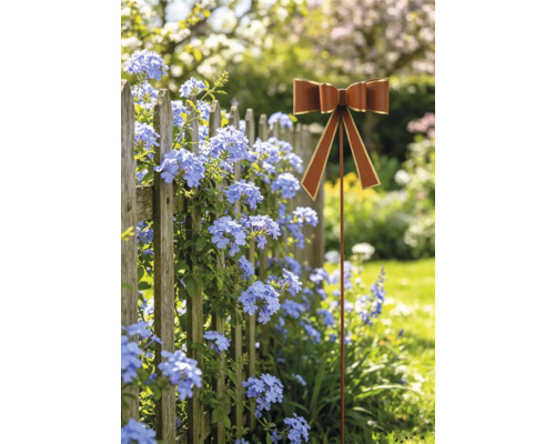 Gartenstecker in Schleifenform in Rostoptik mit goldenem Rand vor einem Holzzaun mit blauen Blumen.