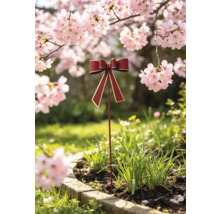 Gartenstecker in Form einer roten Schleife mit goldenem Rand, platziert in einem bepflanzten Beet unter blühenden Kirschbäumen.