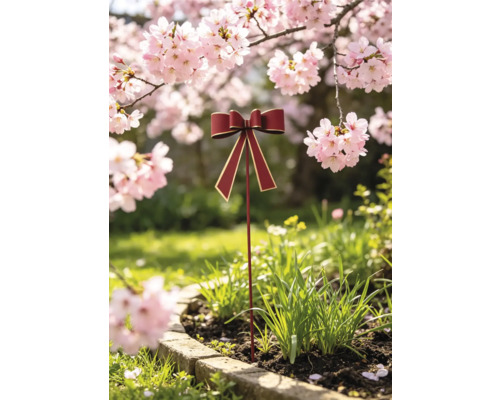 Gartenstecker in Form einer roten Schleife mit goldenem Rand, platziert in einem bepflanzten Beet unter blühenden Kirschbäumen.