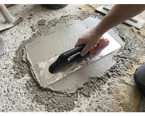 Une main applique du mortier ou de la colle à carrelage sur un sol en béton avec une taloche dentelée.