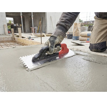 Un artisan utilise une truelle dentelée pour lisser du béton frais sur un chantier.