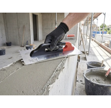 Main avec gant de travail appliquant du mortier sur un mur en béton avec une taloche crantée sur un chantier.