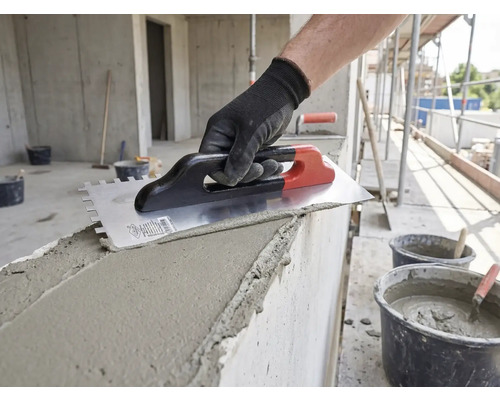 Main avec gant de travail appliquant du mortier sur un mur en béton avec une taloche crantée sur un chantier.