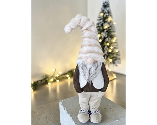 Textiler Weihnachtswichtel mit langer gestreifter Plüschmütze und Bart in einer hellen, festlich dekorierten Umgebung mit Weihnachtsbaum.