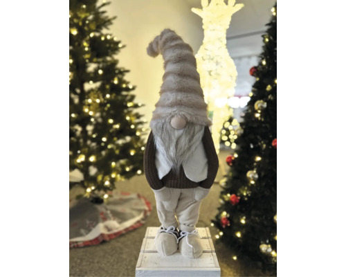 Dekorative Weihnachtswichtel-Figur mit langer Mütze und grauem Bart, stehend zwischen beleuchteten Weihnachtsbäumen.