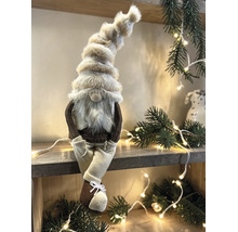 Dekorativer Weihnachtswichtel mit hoher Pelzmütze und Bart sitzend auf einem Holzregal inmitten von Tannenzweigen und einer hellen Lichterkette.