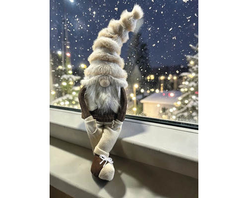 Dekofigur Wichtel mit flauschiger Mütze und weißem Bart, sitzend auf einer Fensterbank vor winterlicher Kulisse mit Lichtern.