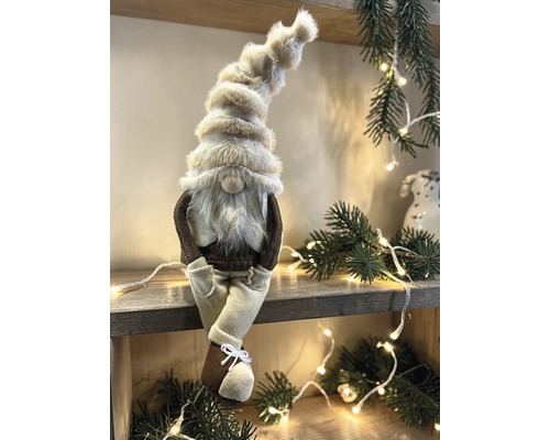 Dekorativer Weihnachtswichtel mit hoher Pelzmütze und Bart sitzend auf einem Holzregal inmitten von Tannenzweigen und einer hellen Lichterkette.