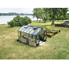 Gewächshaus mit schwarzem Aluminiumrahmen und Ziegelfundament auf Rasenfläche am See, neben hölzernen Hochbeeten.