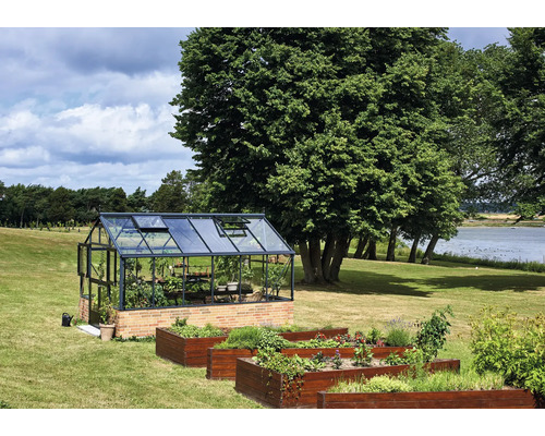 Gewächshaus mit schwarzem Aluminiumrahmen auf einem Ziegelsockel in einem Garten mit Holz-Hochbeeten, Bäumen und einem See im Hintergrund.