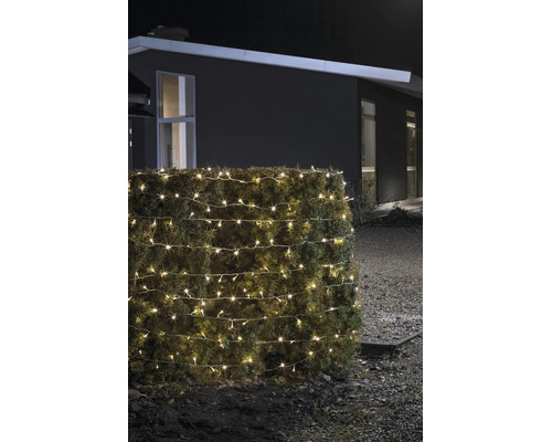Warmweiss leuchtende LED-Lichterkette für den Außenbereich, um eine Hecke gewickelt, bei Nacht vor einem modernen Haus.