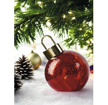 Boule de Noël rouge en verre avec effet craquelé et capuchon doré, sur la neige sous un sapin de Noël illuminé avec des pommes de pin.