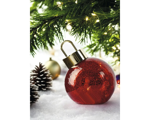 Boule de Noël rouge en verre avec effet craquelé et capuchon doré, sur la neige sous un sapin de Noël illuminé avec des pommes de pin.