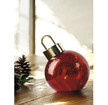 Boule de Noël rouge en verre texturé avec un capuchon doré à côté de pommes de pin sur une surface en bois dans un environnement lumineux.