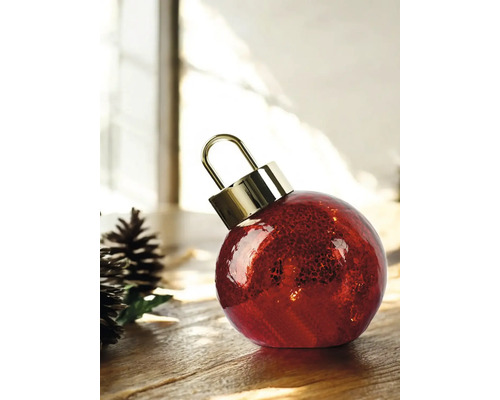 Boule de Noël rouge en verre texturé avec un capuchon doré à côté de pommes de pin sur une surface en bois dans un environnement lumineux.