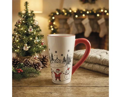 Weisse Keramiktasse mit rotem Henkel, Weihnachtsmann und Schneemann auf Holztisch vor einem beleuchteten Tannenbaum und Kamin.