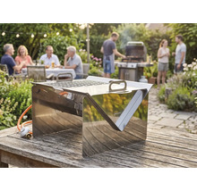 Tisch-Gasgrill aus Edelstahl auf einem Holztisch im Garten, im Hintergrund eine gesellige Runde von Personen beim Grillfest.