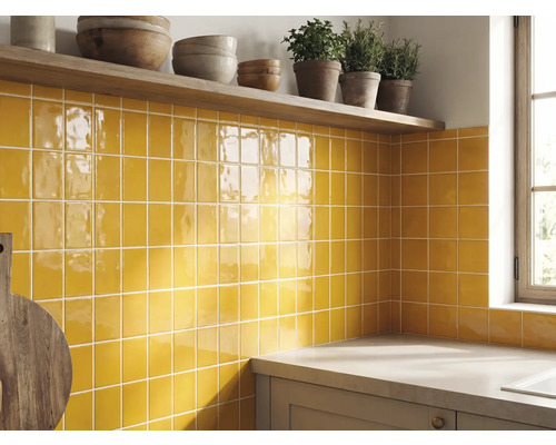 Crédence de cuisine avec carreaux muraux carrés jaunes brillants, une étagère en bois avec des bols et des herbes et un plan de travail.
