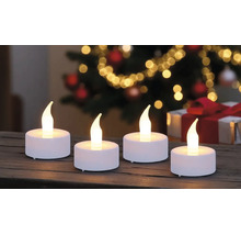 Vier weiße LED-Teelichter mit warm leuchtender Flamme auf einer Holzoberfläche vor einem weihnachtlichen Hintergrund mit Lichtern.