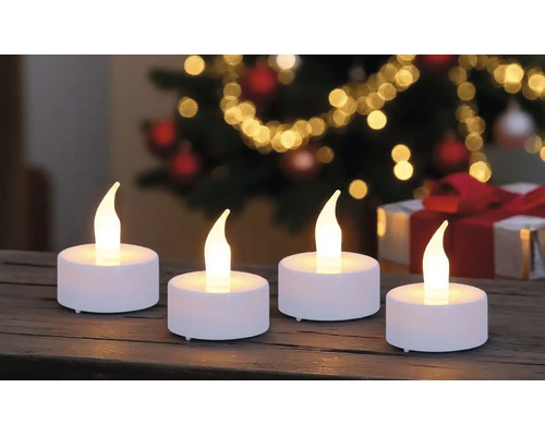 Vier weiße LED-Teelichter mit warm leuchtender Flamme auf einer Holzoberfläche vor einem weihnachtlichen Hintergrund mit Lichtern.