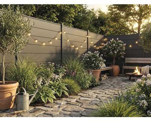 Moderner Garten mit grauem Sichtschutzzaun, Natursteinpflaster, Pflanzen in Terracotta-Töpfen, Lichterkette und brennender Feuerstelle.