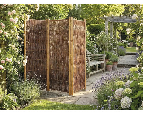 Dreiteiliger faltbarer Sichtschutz aus Weide in einem Garten mit Steinplatten, Blumen und einer Gartenbank.