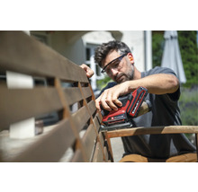 Ein Mann schleift eine Gartenbank aus Holz mit einem batteriebetriebenen Einhell Multischleifer im Freien und trägt dabei eine Schutzbrille.