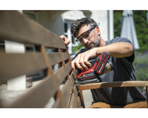 Ein Mann schleift eine Gartenbank aus Holz mit einem batteriebetriebenen Einhell Multischleifer im Freien und trägt dabei eine Schutzbrille.