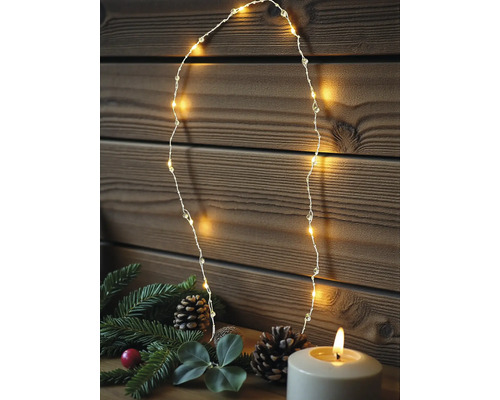 Warmweiße LED-Drahtlichterkette vor einer Holzwand, dekoriert mit Tannenzweigen, Zapfen und einer brennenden Kerze.