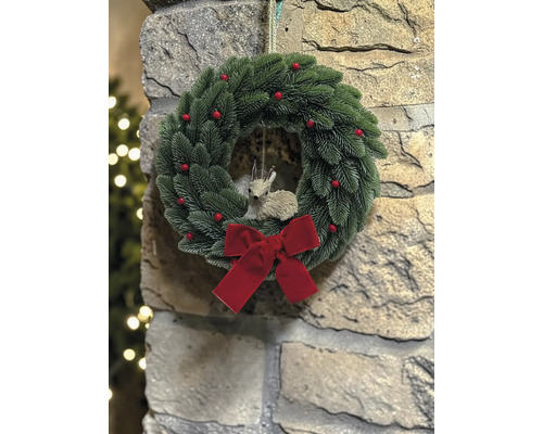 Weihnachtskranz mit Tannennadeln, roten Beeren, einer roten Schleife und einer Rehfigur an einer Steinwand.