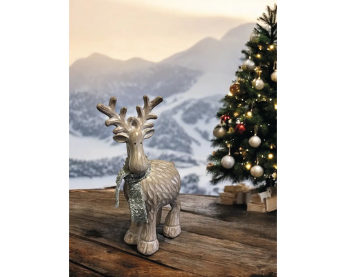 Rentier-Figur mit silbernem Glitzerschal vor einem geschmückten Weihnachtsbaum und Bergkulisse auf einem Holztisch.