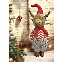 Weihnachtsfigur Highland-Rind mit roter Mütze, Schal und gestreifter Kleidung neben einem Weihnachtsbaum vor einer Steinwand.