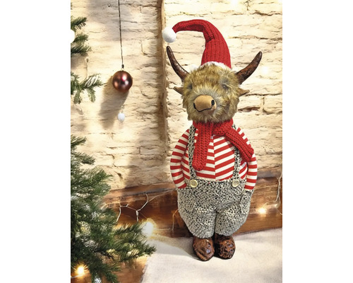 Weihnachtsfigur Highland-Rind mit roter Mütze, Schal und gestreifter Kleidung neben einem Weihnachtsbaum vor einer Steinwand.
