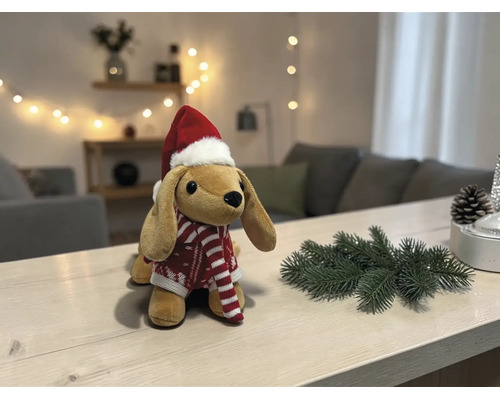 Plüschhund mit Weihnachtsmütze und rot-weiß gestreiftem Pullover auf einem hellen Holztisch mit Tannenzweig und Tannenzapfen.