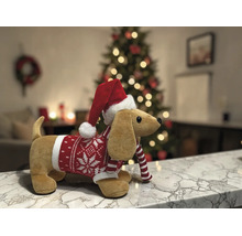 Plüsch-Dackel mit Weihnachtsmütze und Strickpullover auf einer Marmoroberfläche vor einem beleuchteten Weihnachtsbaum in einem hellen Raum.