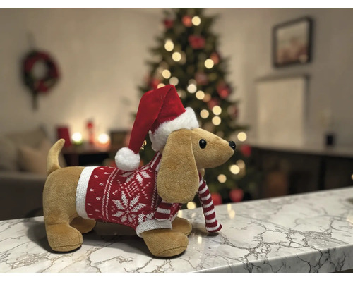 Plüsch-Dackel mit Weihnachtsmütze und Strickpullover auf einer Marmoroberfläche vor einem beleuchteten Weihnachtsbaum in einem hellen Raum.