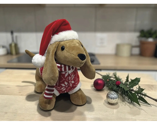 Stofftier Dackel mit Weihnachtsmütze und Strickpullover auf einem Holztisch neben Weihnachtsdekoration.