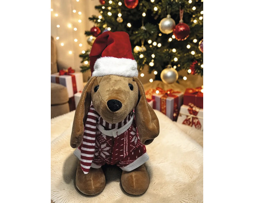 Stoffhund mit Weihnachtsmütze, gestreiftem Schal und Pullover vor einem geschmückten Weihnachtsbaum mit Geschenken.