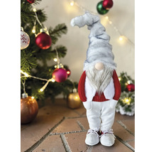 Weihnachtsgnom mit grauer Mütze, weißem Bart, rotem Oberteil und weißer Hose vor einem geschmückten Tannenbaum mit Lichtern.