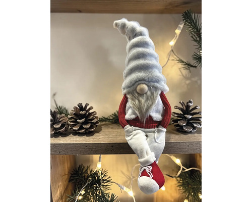 Weihnachts-Wichtel Figur mit weißer Zipfelmütze, langem Bart, rotem Pullover und weißer Hose sitzend auf einem Holzregal.