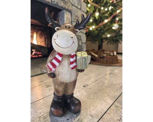 Weihnachtsdekoration Rentier-Figur mit gestreiftem Schal und Geschenk vor Kamin und Tannenbaum.