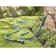 Textiler Gartenschlauch angeschlossen an einen Außenwasserhahn an einer Ziegelwand, liegend auf einer Rasenfläche im Garten.