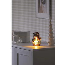 Décoration de Noël ours avec sapin lumineux, écharpe et bonnet de Père Noël sur une commode blanche à côté d'un cadeau et d'un sapin décoratif.