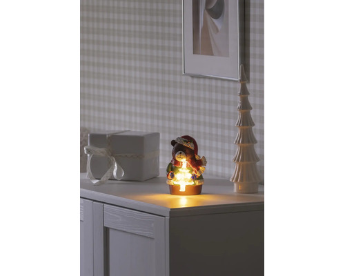 Décoration de Noël ours avec sapin lumineux, écharpe et bonnet de Père Noël sur une commode blanche à côté d'un cadeau et d'un sapin décoratif.