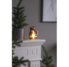 Figurine de Noël ours avec éclairage LED tenant un petit sapin lumineux, posée sur un manteau de cheminée blanc à côté d'un sapin en pot.