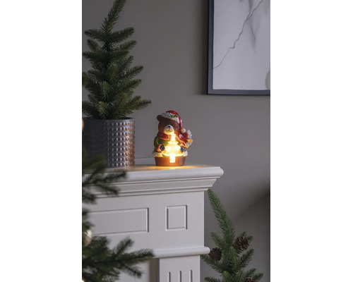 Figurine de Noël ours avec éclairage LED tenant un petit sapin lumineux, posée sur un manteau de cheminée blanc à côté d'un sapin en pot.