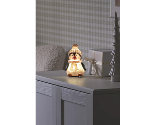Figurine décorative LED pingouin avec bonnet et écharpe sur un buffet à côté d'un cadeau et d'un arbre de décoration.