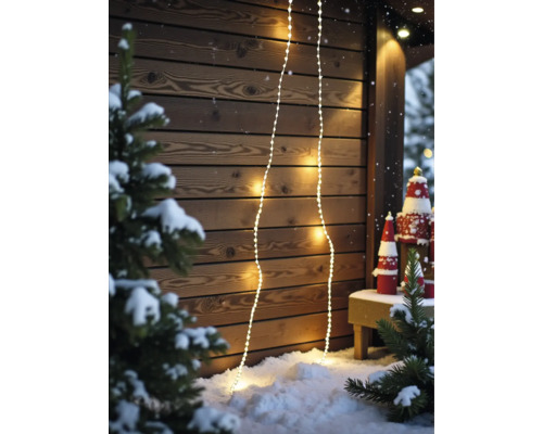 Warmweisse LED-Lichterketten an einer hölzernen Wand in einer verschneiten Szene mit Tannenbäumen und Weihnachtsdekoration.