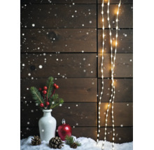 Weihnachtliche Dekoration mit LED-Lichterketten vor einer Holzwand, Vase mit Tannenzweigen, Beeren, Zapfen und einer Weihnachtskugel im Schnee.